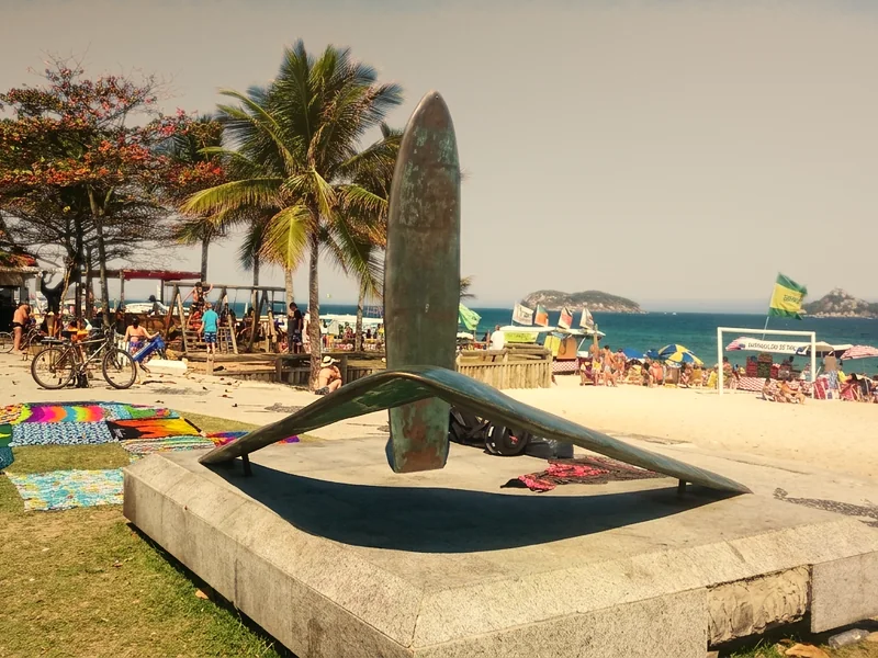 Escultura de prancha e asa-delta na orla da Praia do Pepê, com coqueiros e o mar ao fundo